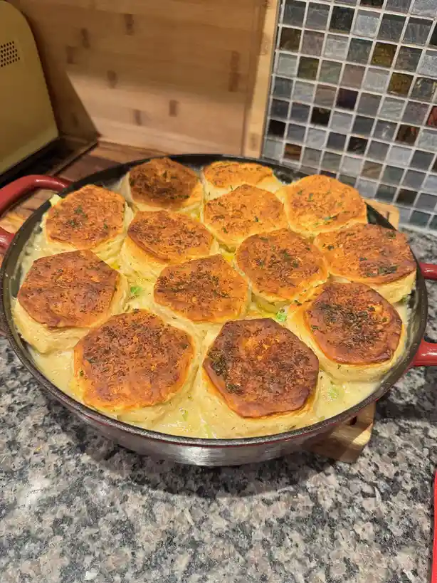 A close-up photo shows a red-handled cast iron skillet filled with a creamy dish topped with golden-brown, baked biscuits…