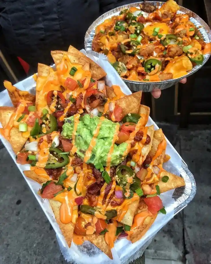 Two hands hold up aluminum trays filled with heavily loaded nachos, topped with melted cheese, guacamole, jalapeños…