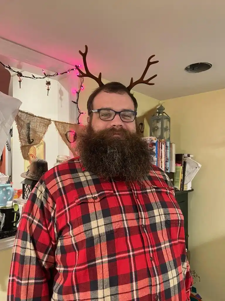 a man with a thick beard and glasses wears a red plaid shirt and brown plastic reindeer antlers on his head. The background…