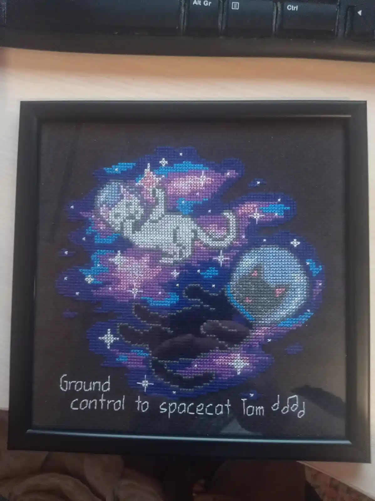 A framed cross-stitch artwork depicts a cat in space, surrounded by stars and nebulae in purple and blue hues, with the text…