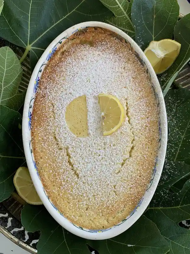 An overhead shot of a golden-brown lemon cake in an oval ceramic dish, dusted with powdered sugar to form a smiling face…