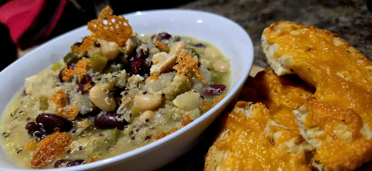 A close-up photo shows a white bowl filled with a hearty bean and quinoa stew topped with crispy fried wonton strips, next…