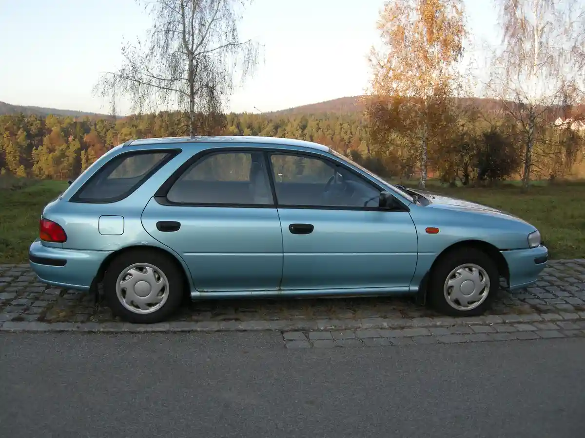 A light blue 1990s-era hatchback car is parked on a cobblestone curb beside a road, with a forested hillside and autumn…