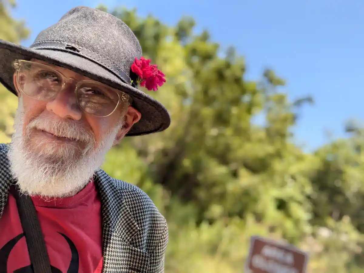 An older man with a white beard and glasses smiles at the camera while wearing a grey hat adorned with a bright pink flower…