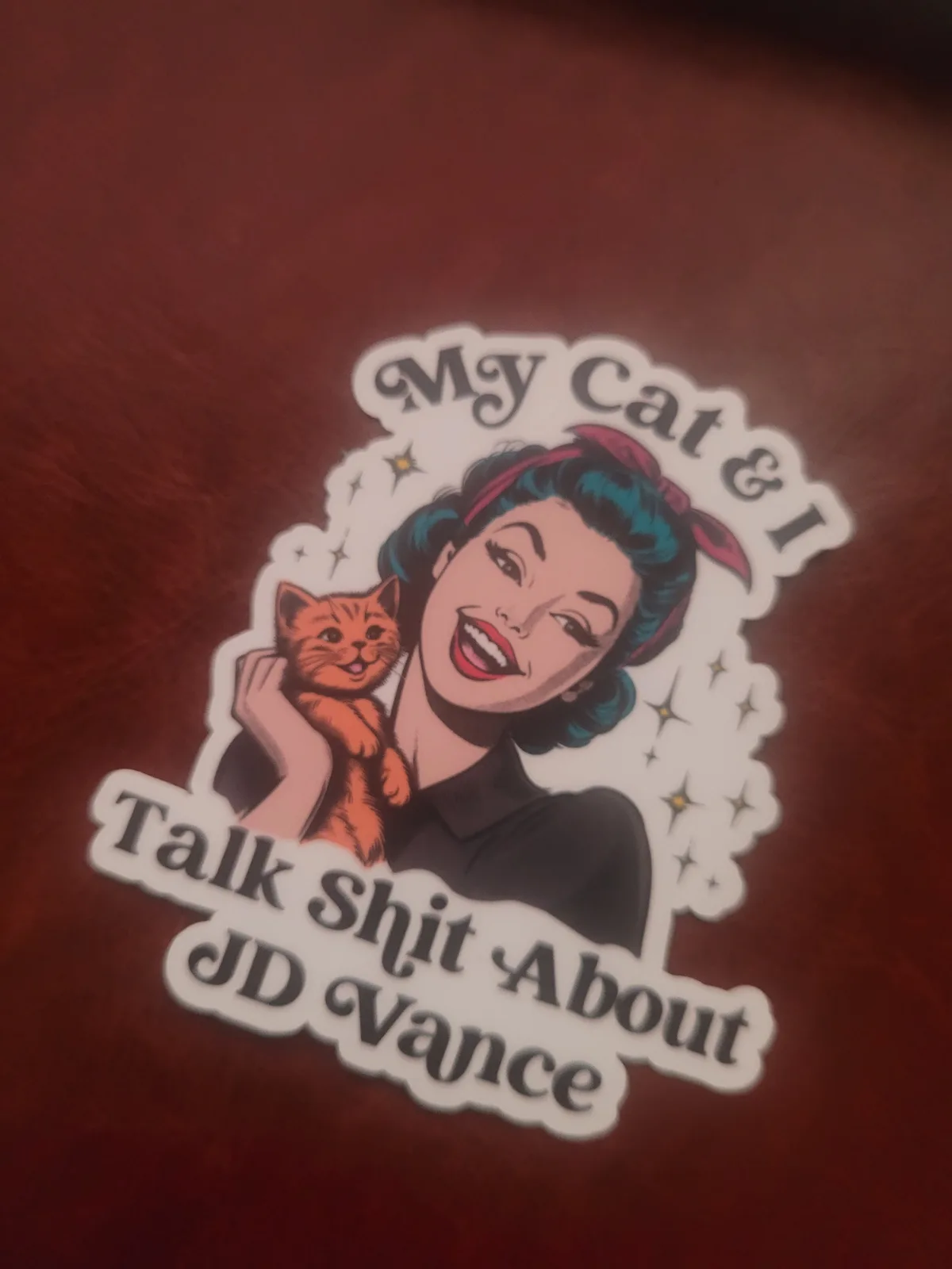 A sticker features a vintage-style illustration of a smiling woman with blue hair and a red headband holding an orange cat…