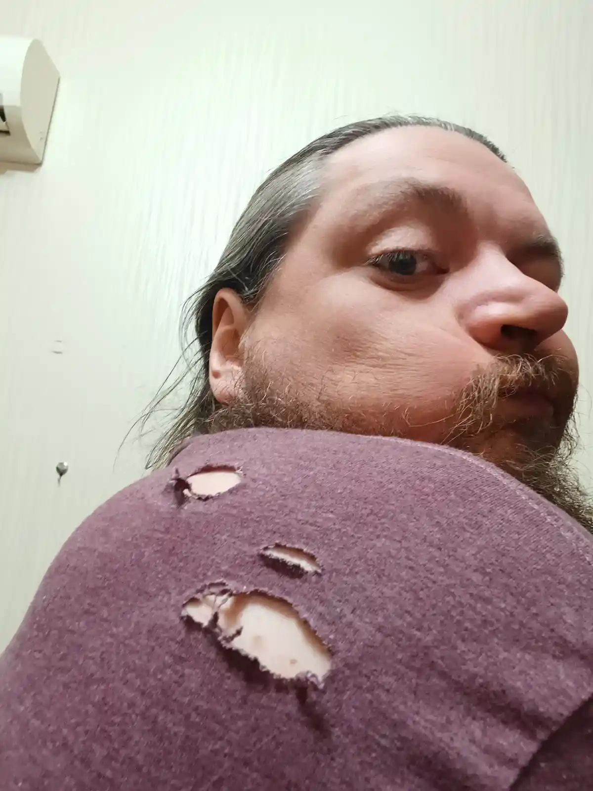 A man with long hair and a beard looks at the camera while wearing a purple shirt with multiple large, ragged holes in the…