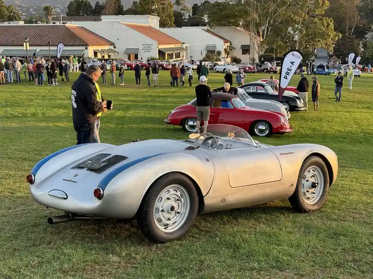 A silver Porsche Spyder race car with blue stripes is parked on grass at a classic car show, with people and other vintage…