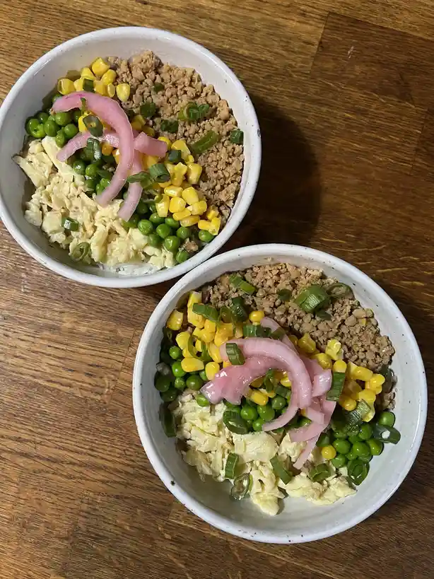 Two white bowls sit on a wooden table, each filled with rice topped with scrambled eggs, ground meat, green peas, yellow…
