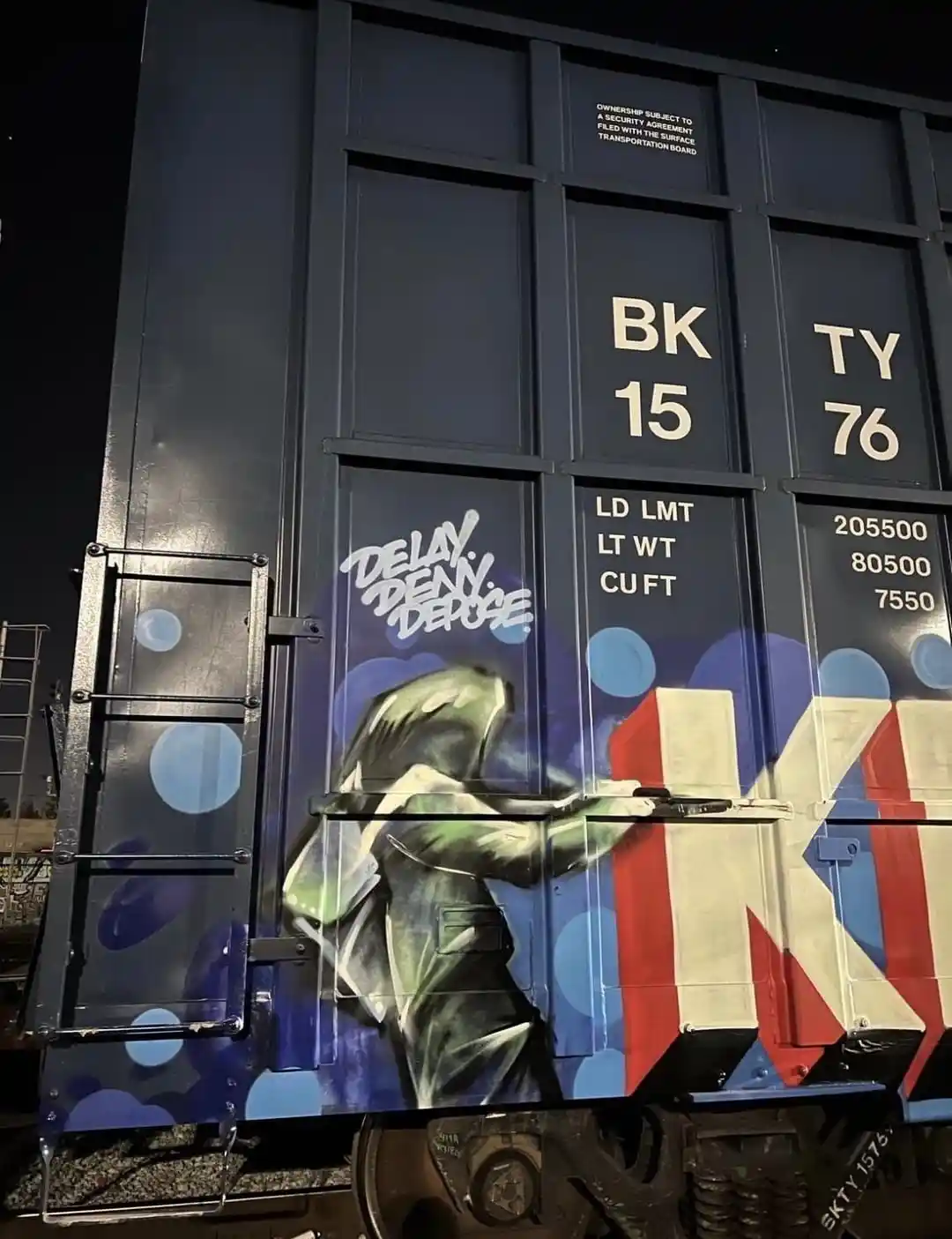 A nighttime photo of a dark blue freight train car with graffiti art showing a green figure in a suit holding a large…