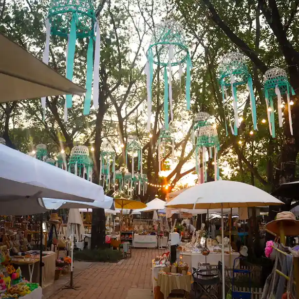 An outdoor market scene at dusk with numerous teal and white woven lanterns hanging from trees, string lights, and vendor…