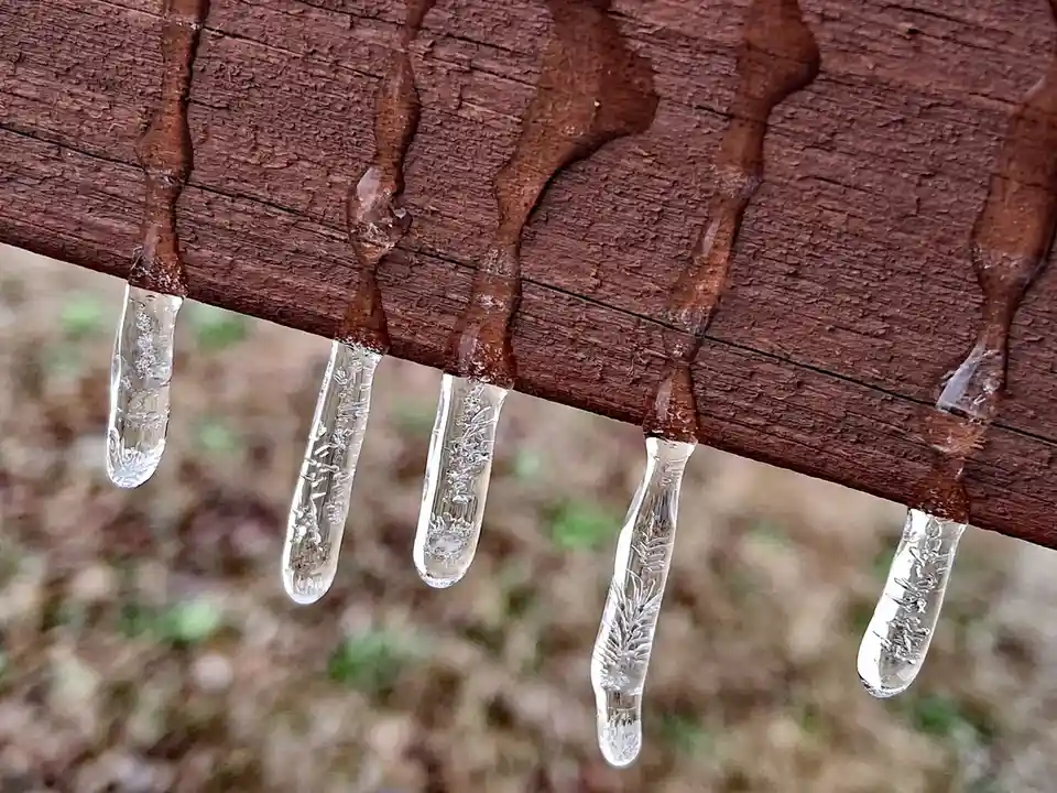 A close-up photo shows several clear, elongated icicles hanging from the edge of a weathered, brown wooden surface. The…