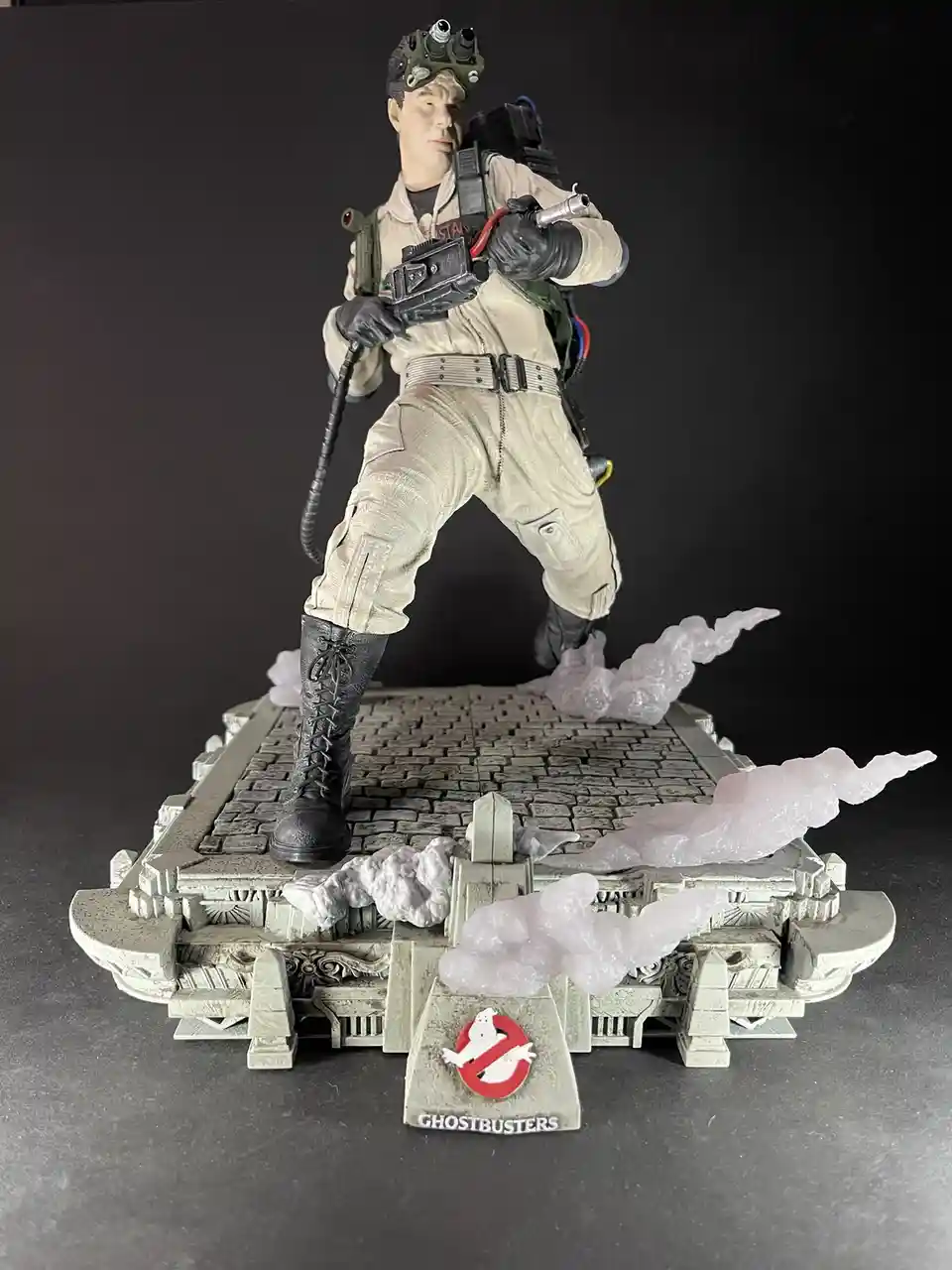 A Ghostbusters action figure of the character Peter Venkman is posed on a detailed diorama base featuring ghostly smoke…