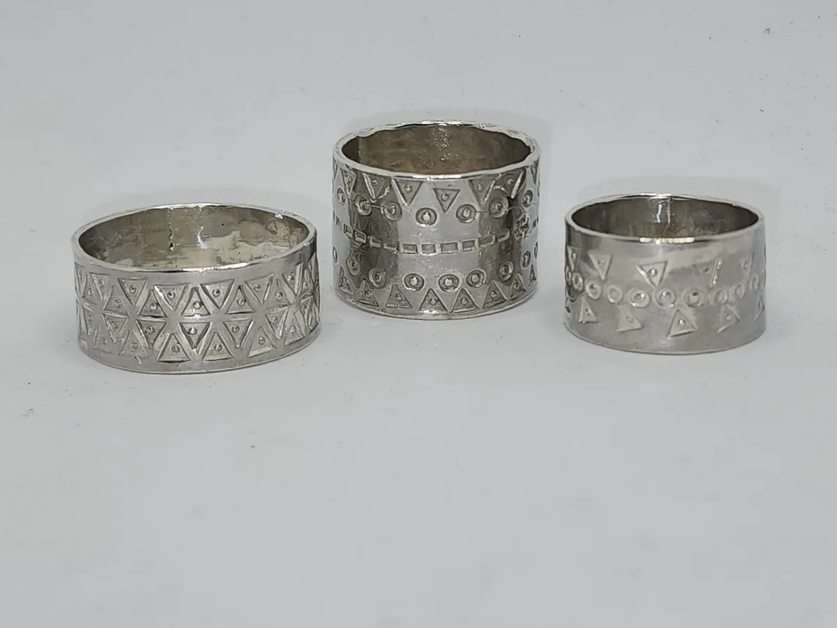 Three silver-colored napkin rings with geometric patterns sit on a plain white surface. The middle ring is noticeably taller…