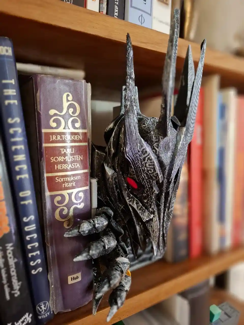 A dark, menacing Sauron mask with glowing red eyes and sharp horns is perched on a wooden bookshelf, gripping a purple book…