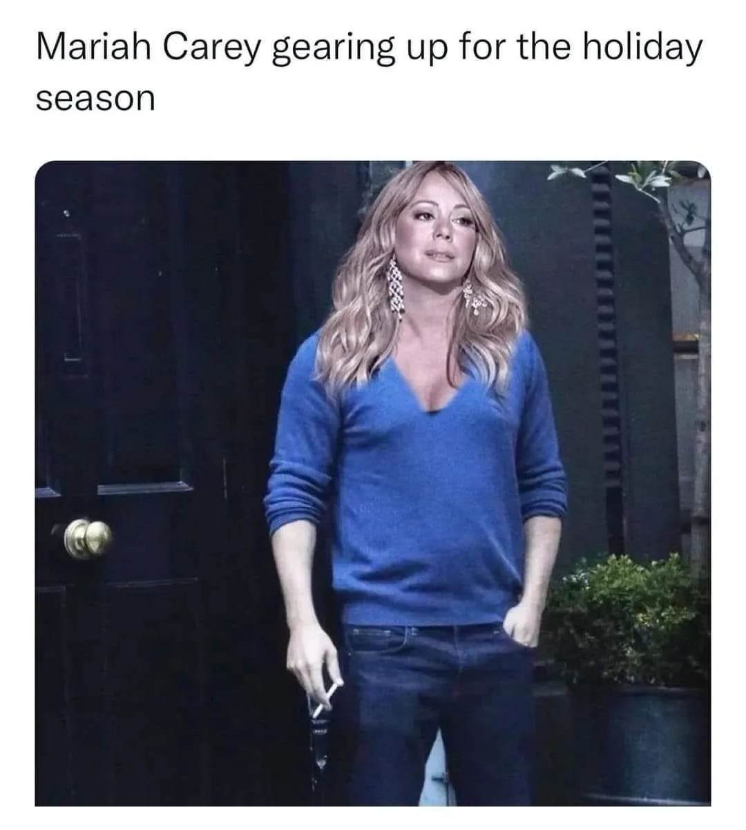 A woman with blonde hair wearing a blue v-neck sweater and jeans stands in front of a dark door, holding a cigarette. The…
