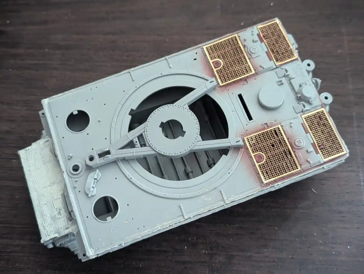 A partially assembled grey plastic model tank hull with gold-colored grilles and visible internal components rests on a dark…