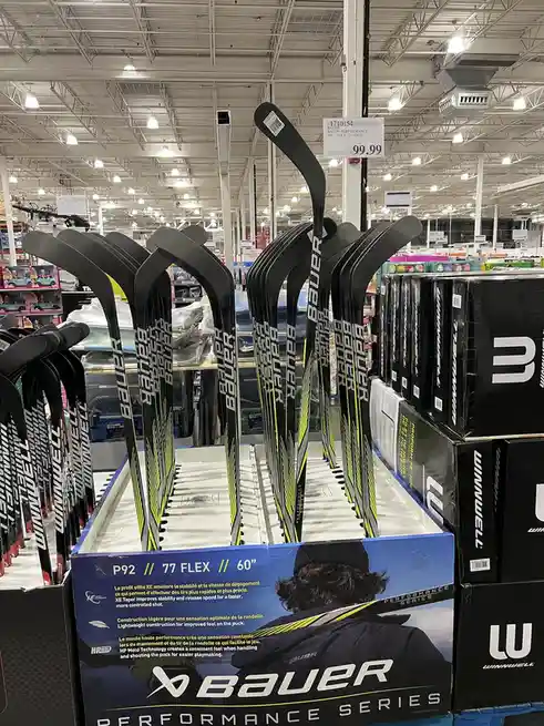 A display of Bauer hockey sticks in a warehouse store, priced at $99.99, with the price tag clearly visible above the…