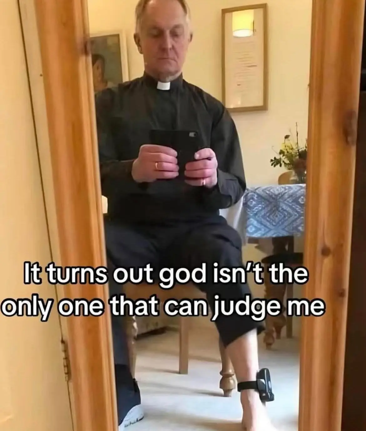 A man in a black clerical collar shirt is sitting on a wooden chair, looking down at his phone. He has a black ankle monitor…