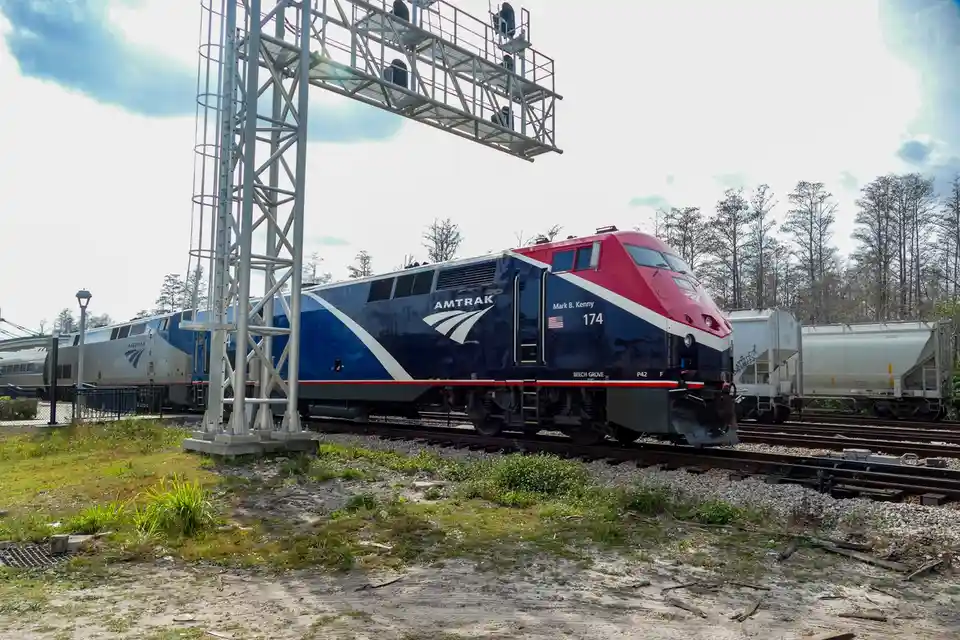 An Amtrak locomotive, numbered 174 and named 'Mark B. Kenny', is shown on train tracks with a large metal signal gantry…