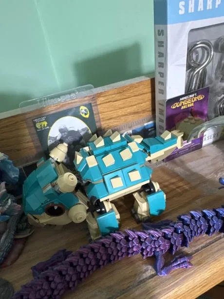 A teal and beige LEGO-style dinosaur model sits on a wooden surface next to a purple plastic dinosaur spine. Behind it are…