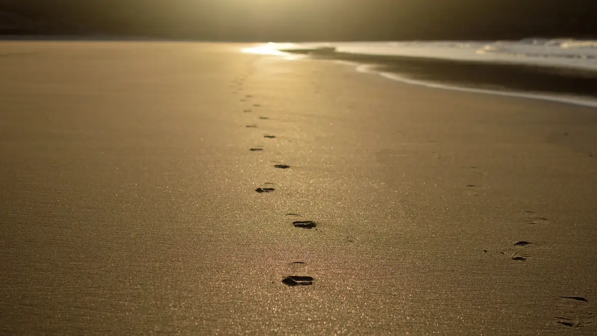 A low-angle shot of a sandy beach at sunset, with a clear trail of footprints leading from the foreground toward the ocean…