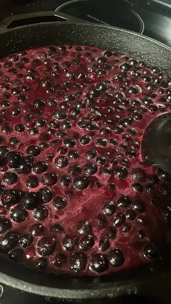 A close-up photo shows a black cast-iron skillet filled with dark purple blueberries simmering in their own juice on a…