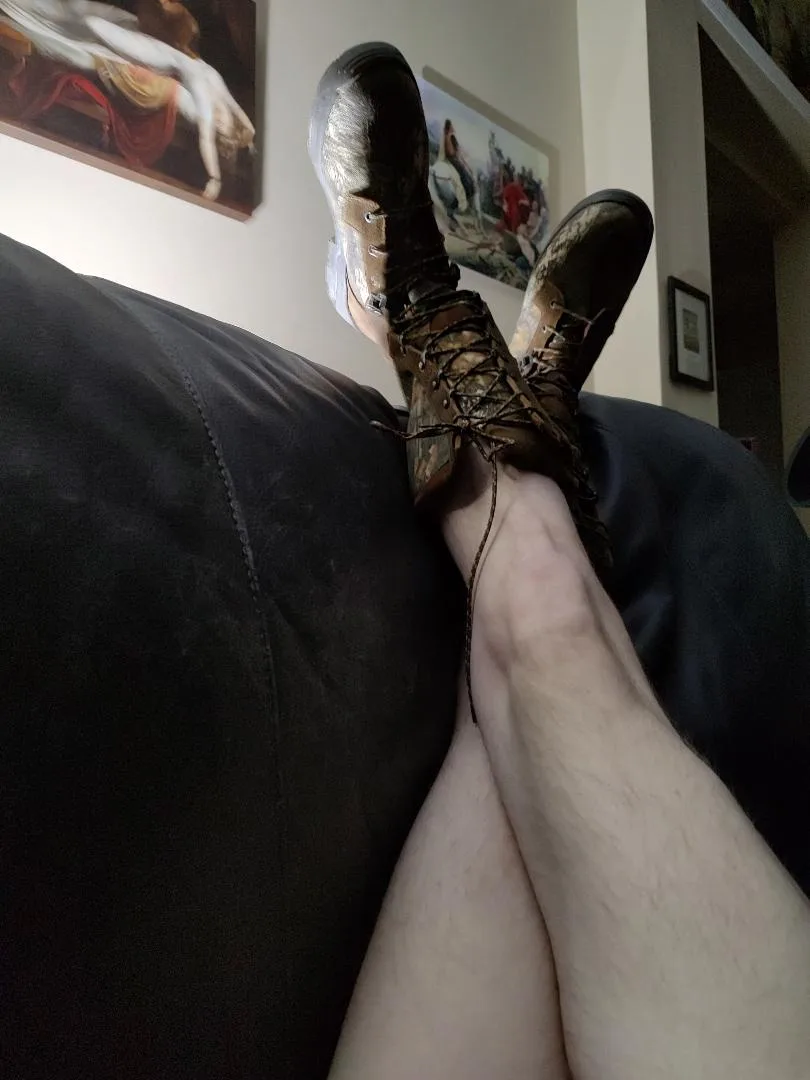 A person is lounging on a dark couch with their legs propped up, wearing muddy, laced hiking boots. Behind them, two framed…