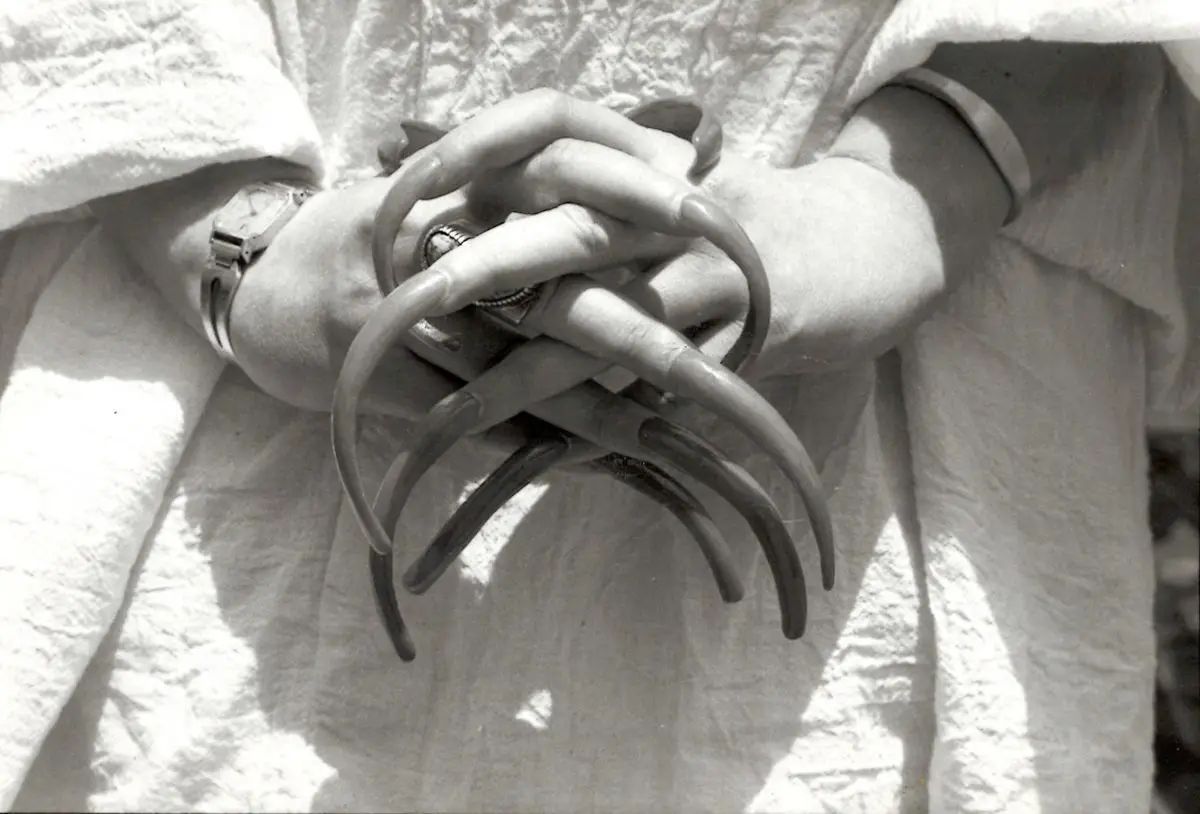 A black-and-white photo shows a person's hands clasped together, with long, curved, claw-like fingernails. The person wears…