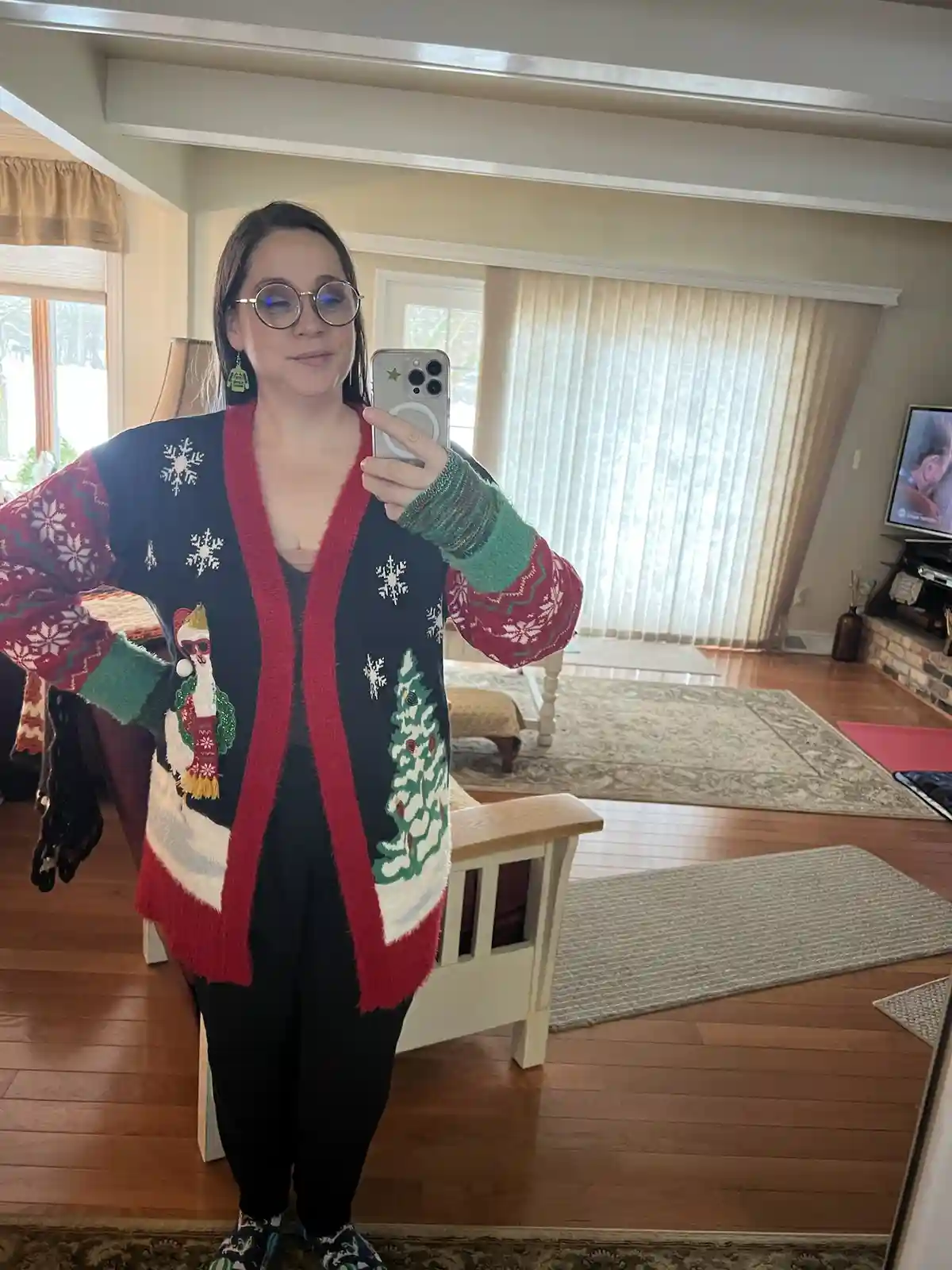 A woman takes a mirror selfie in a very busy, colorful Christmas sweater featuring snowflakes, a penguin in a Santa hat, and…