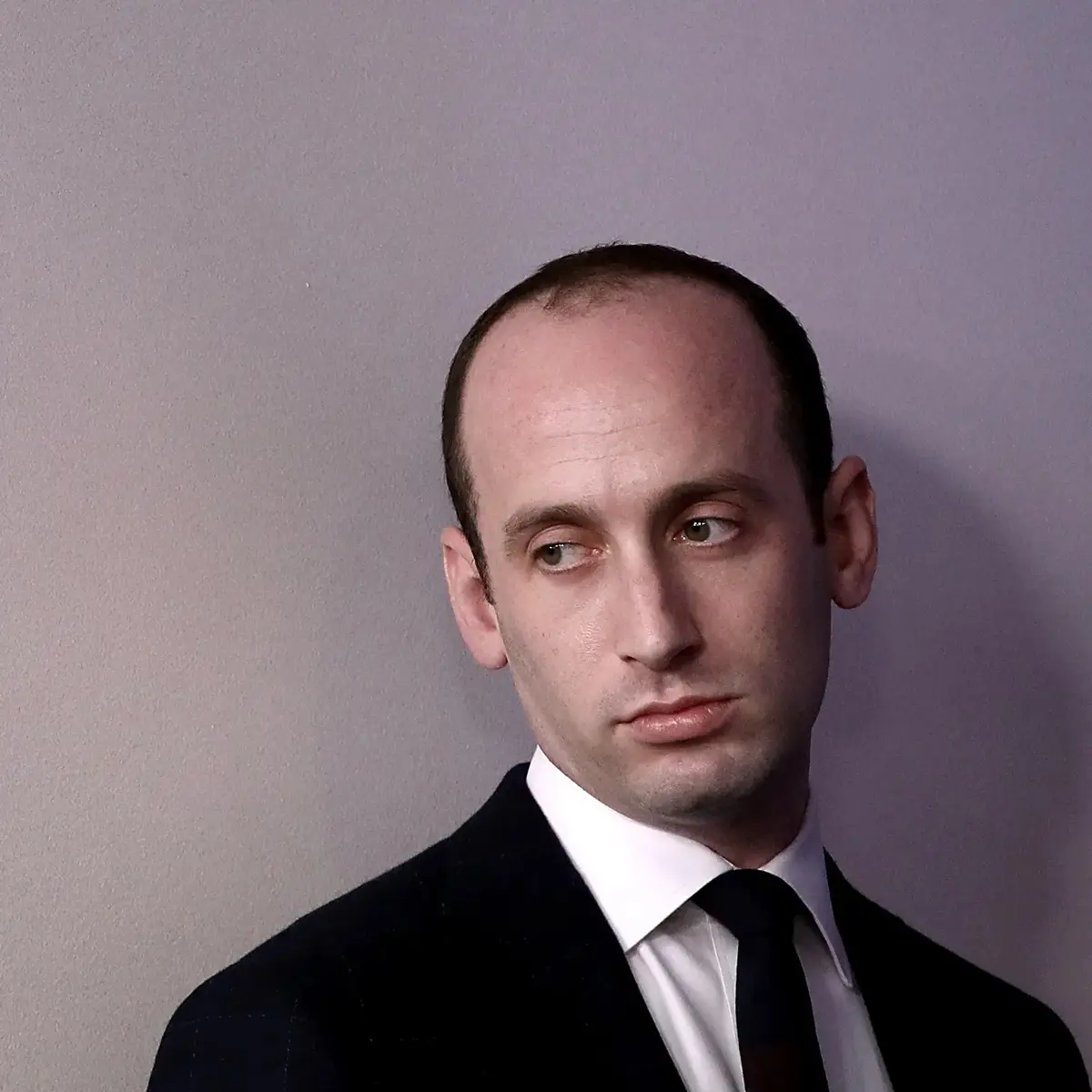 A man in a dark suit and tie stands against a plain gray wall, looking sideways with a skeptical, unimpressed expression…