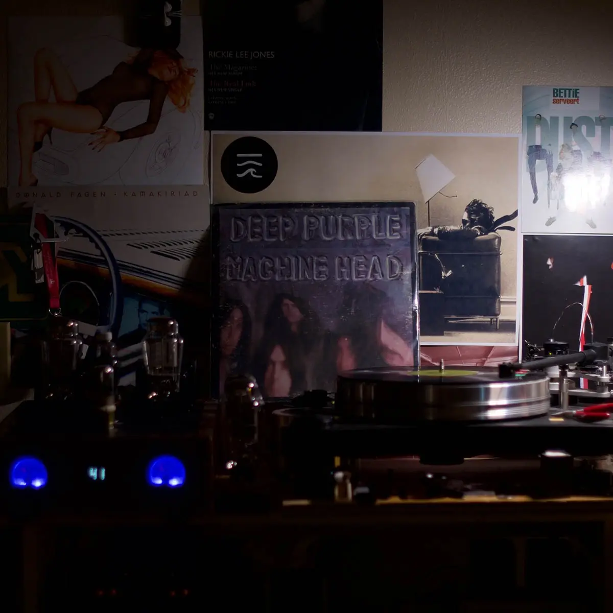 A dimly lit room features a record player with a vinyl on the turntable, surrounded by various album covers including 'Deep…
