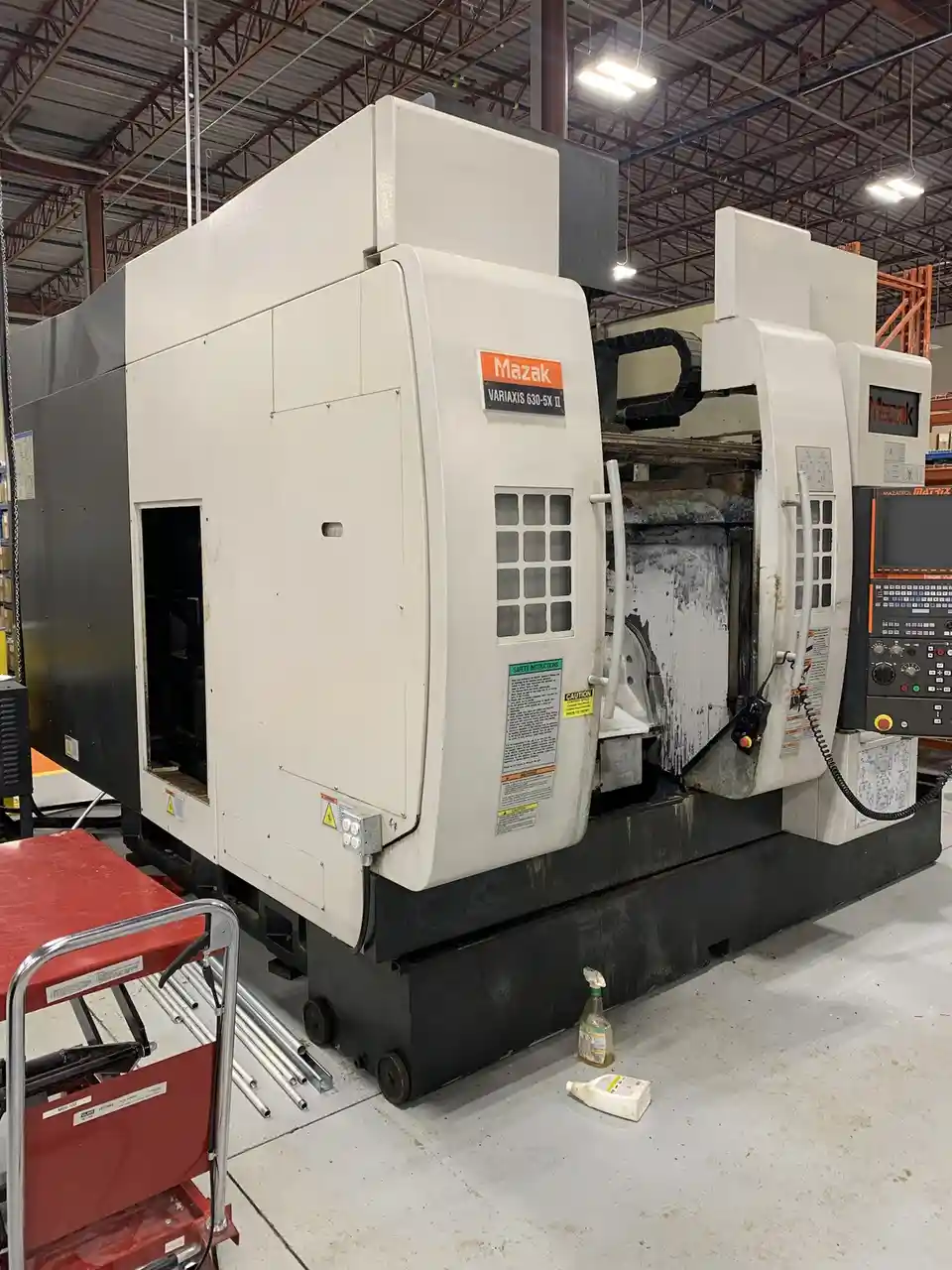 A large, white and black Mazak Variaxis 630-5X II CNC machine sits in a warehouse, visibly dirty with oil stains on its open…