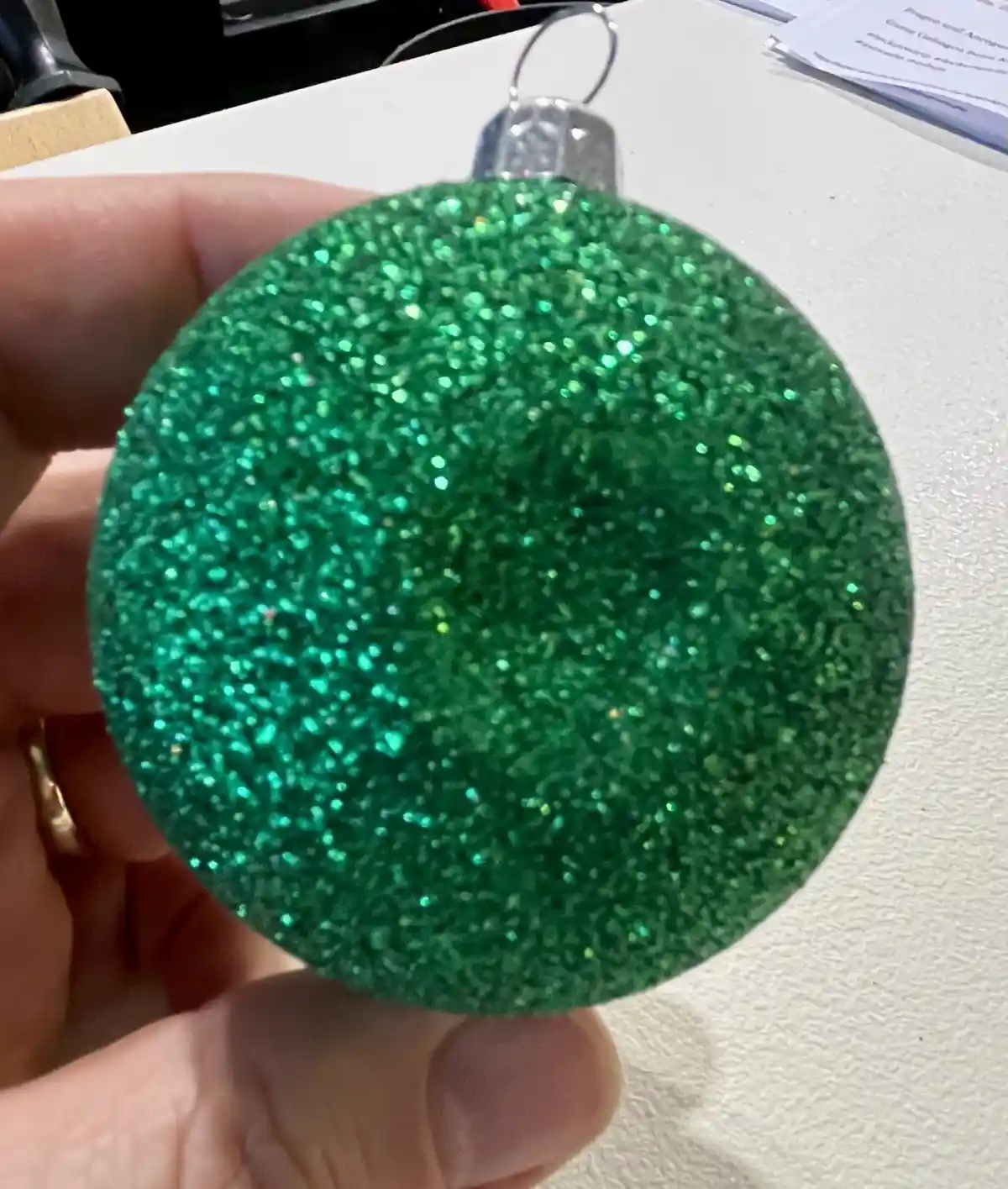 A hand holds a round, green glitter-covered Christmas ornament with a silver cap and loop, against a white surface. The…