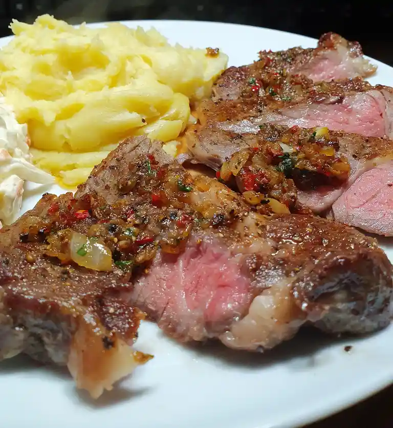 A close-up photo of a plate of sliced medium-rare steak topped with a savory onion and pepper relish, served with creamy…