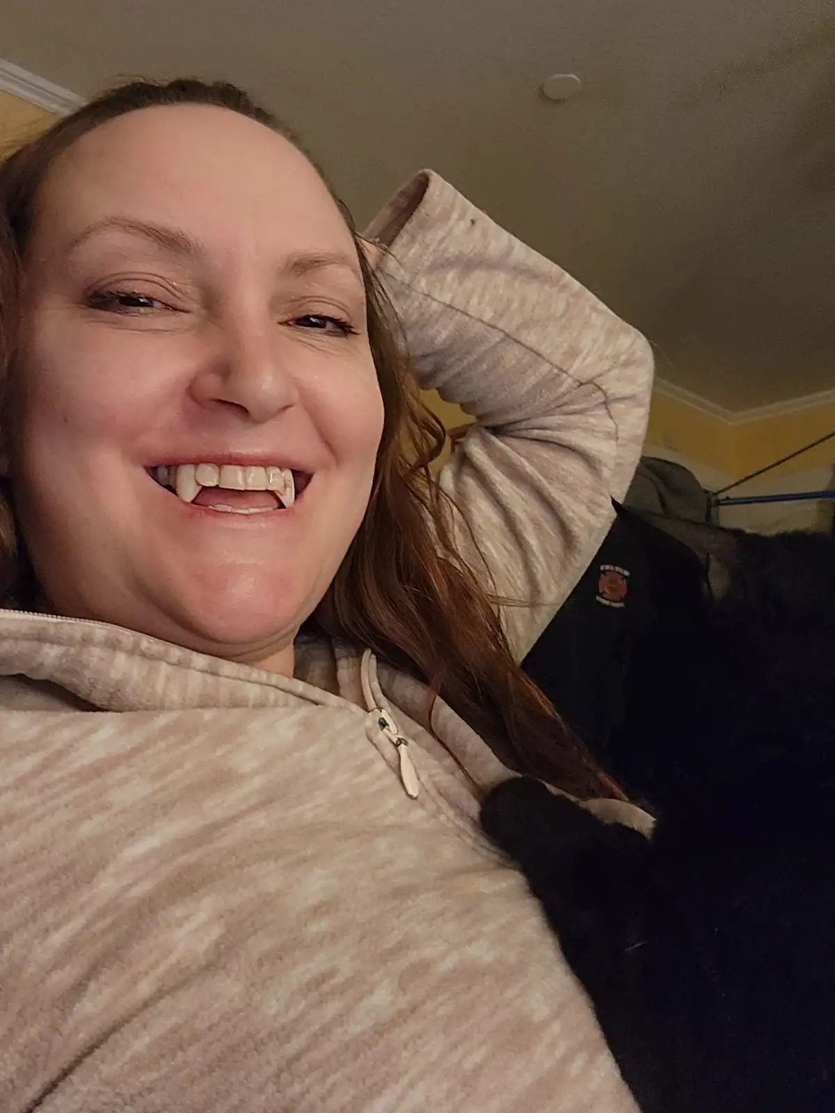 A woman with long brown hair, wearing a beige zip-up hoodie, smiles widely at the camera while lying down with her hand…