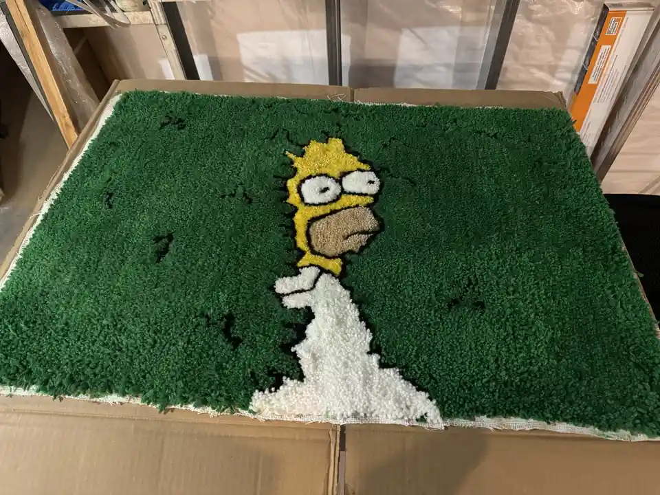 A handmade, shaggy green rug depicts Homer Simpson in a side profile, rendered in textured yarn with yellow hair, white…