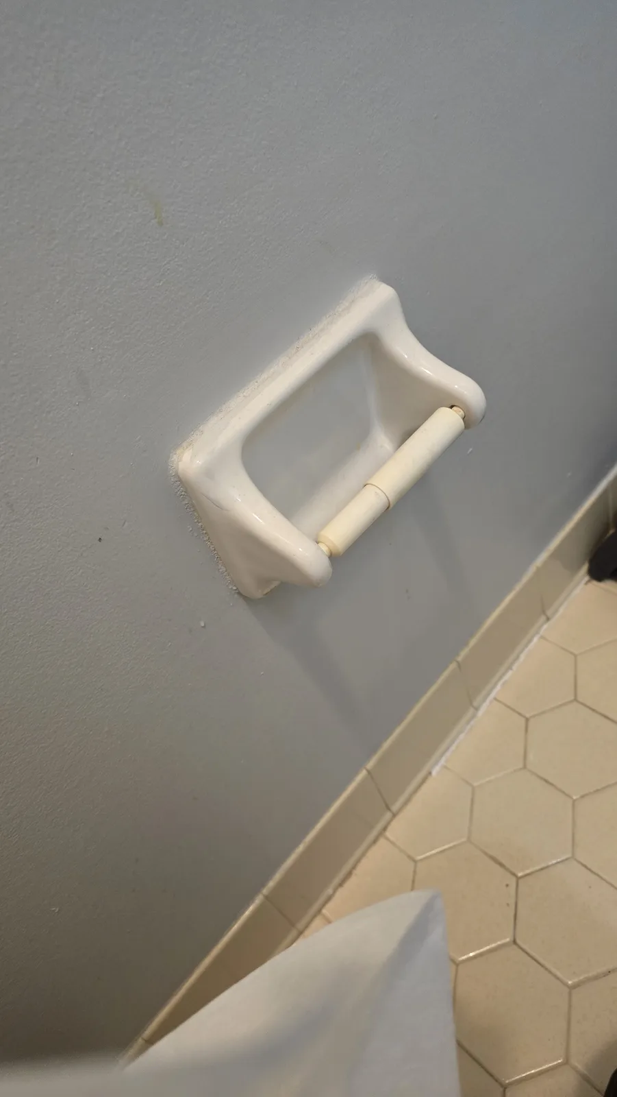 A close-up photo shows an empty white ceramic toilet paper holder mounted on a gray wall, with beige hexagonal tiles visible…
