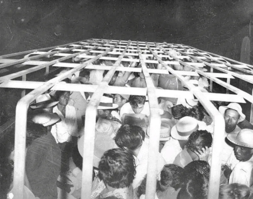 A black-and-white historical photo shows a large crowd of people packed tightly inside a metal cage structure, viewed from…