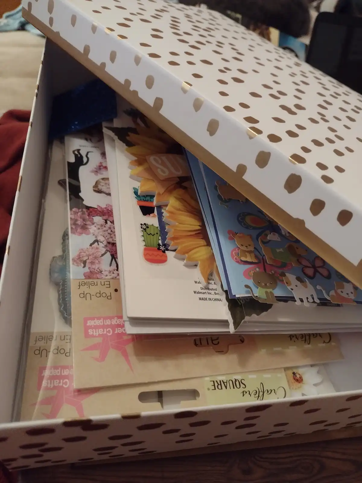A white cardboard box with a gold polka-dot pattern is open, revealing stacks of colorful sticker sheets inside. The visible…