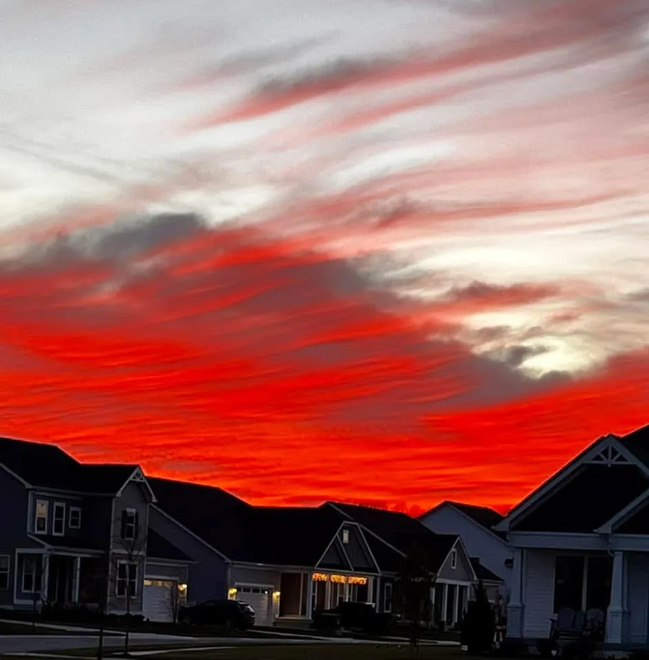 A dramatic, fiery red-orange sunset with streaky clouds dominates the sky above a row of suburban houses, whose silhouettes…