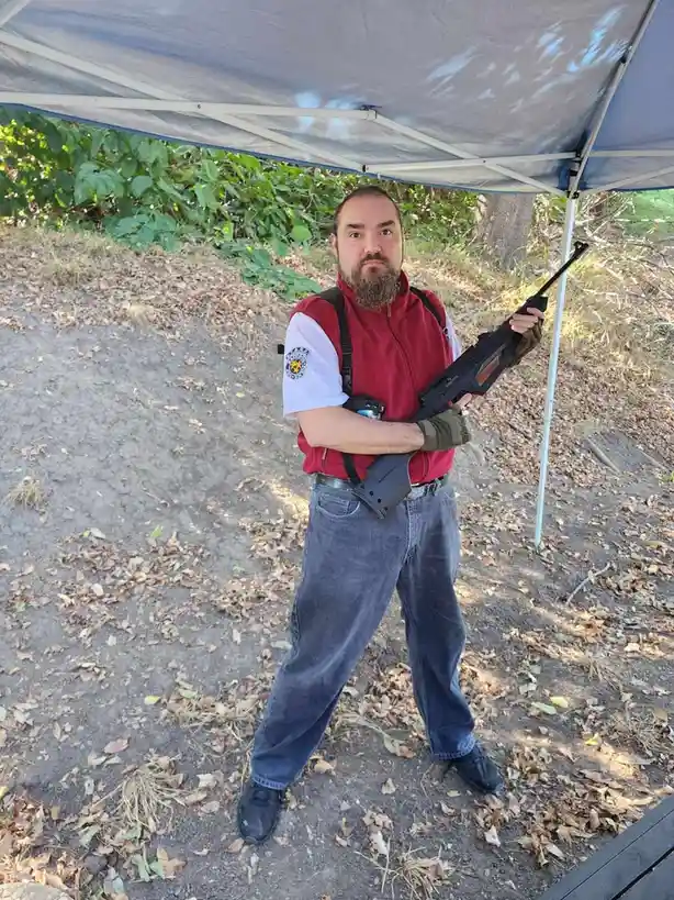A man with a beard and mustache stands under a blue pop-up tent outdoors, holding a black airsoft rifle with both hands. He…