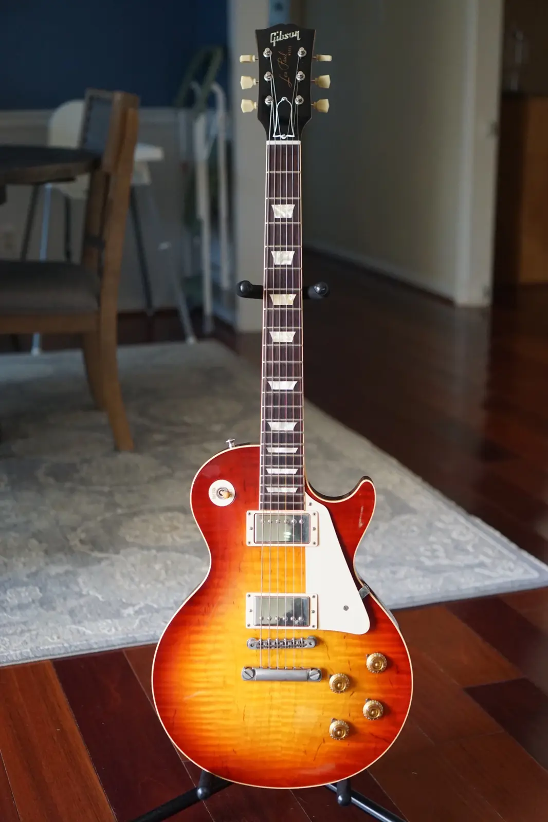 A Gibson Les Paul electric guitar with a sunburst finish stands upright on a black stand in a room with hardwood floors and…