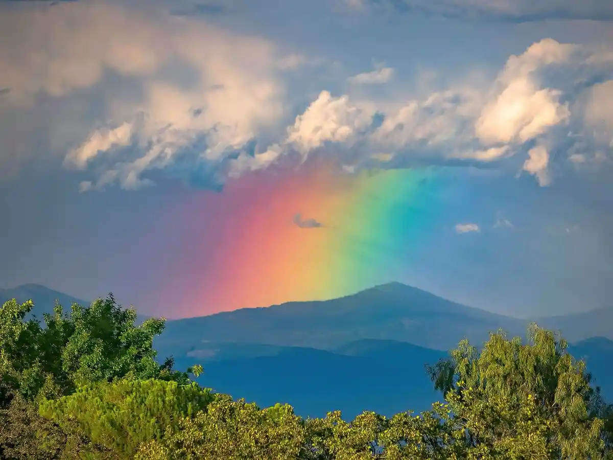 A scenic landscape photo shows a vibrant rainbow arcing across a partly cloudy sky above distant blue mountains, framed by…