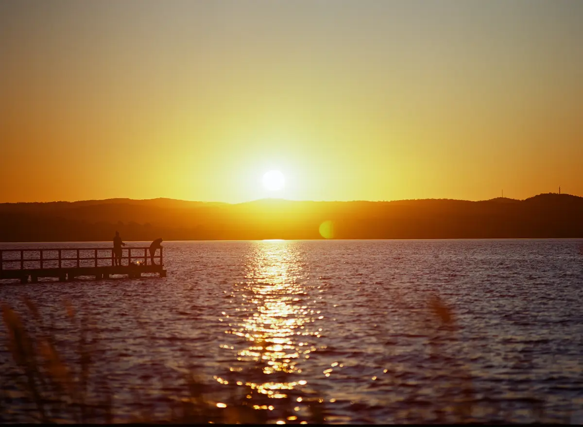 A serene sunset over a lake with a wooden pier extending into the water. Two silhouetted figures stand on the pier, one…