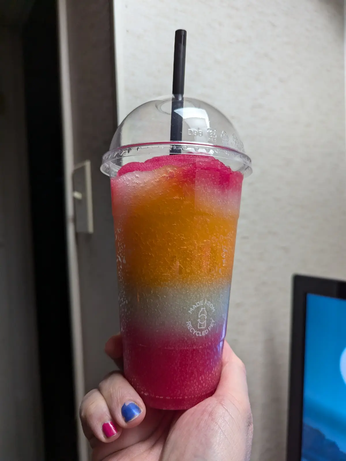 A hand holds a clear plastic cup with a dome lid and black straw, filled with a vibrant, layered frozen drink that…