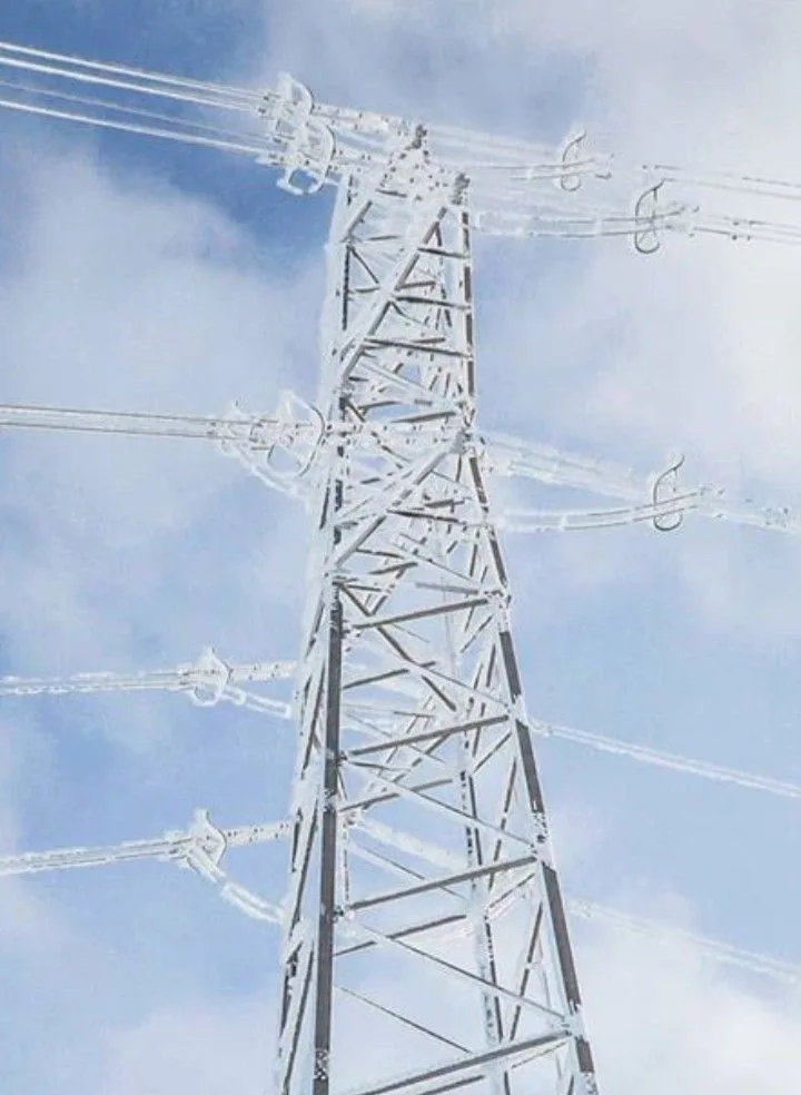 A low-angle photo of a tall, white-painted electricity pylon against a partly cloudy blue sky, with thick ice coating the…