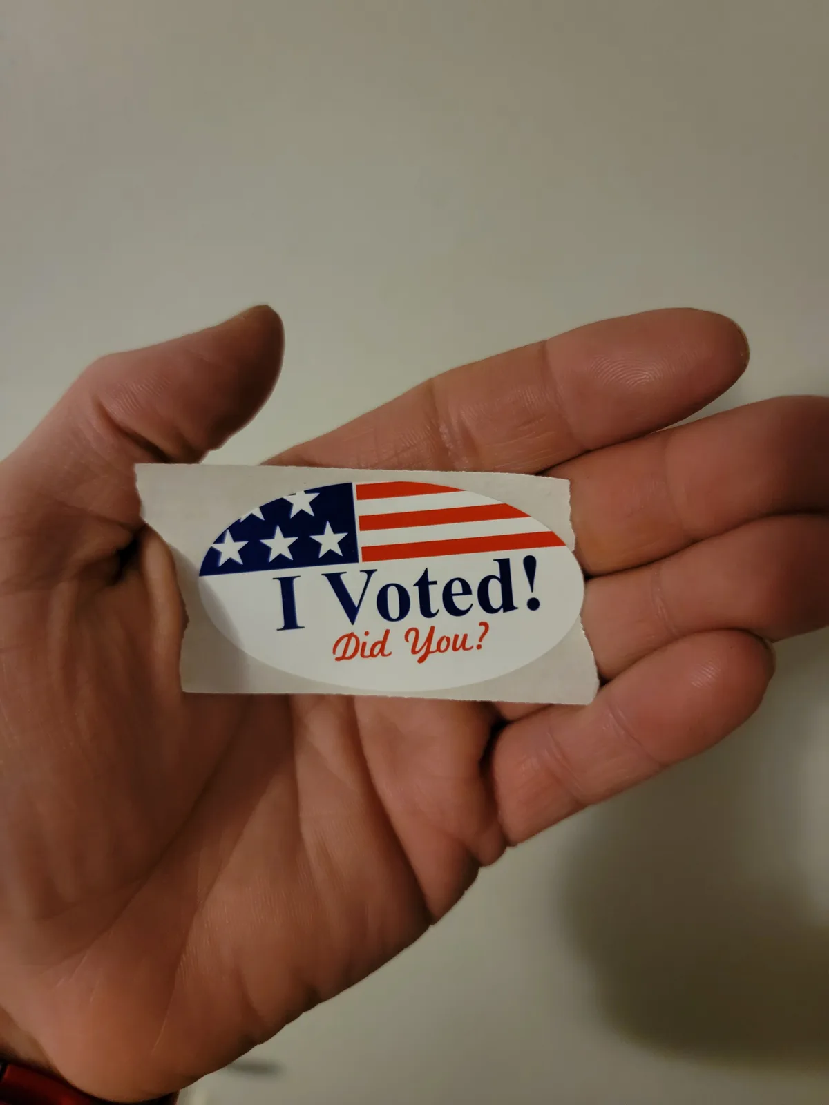 A hand holds a torn 'I Voted!' sticker featuring a half American flag design with stars and stripes. The sticker has bold…