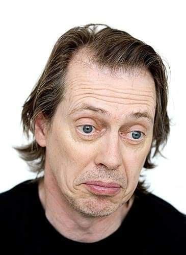 A man with medium-length brown hair and blue eyes stares with wide, startled eyes and a slightly downturned mouth, wearing a…