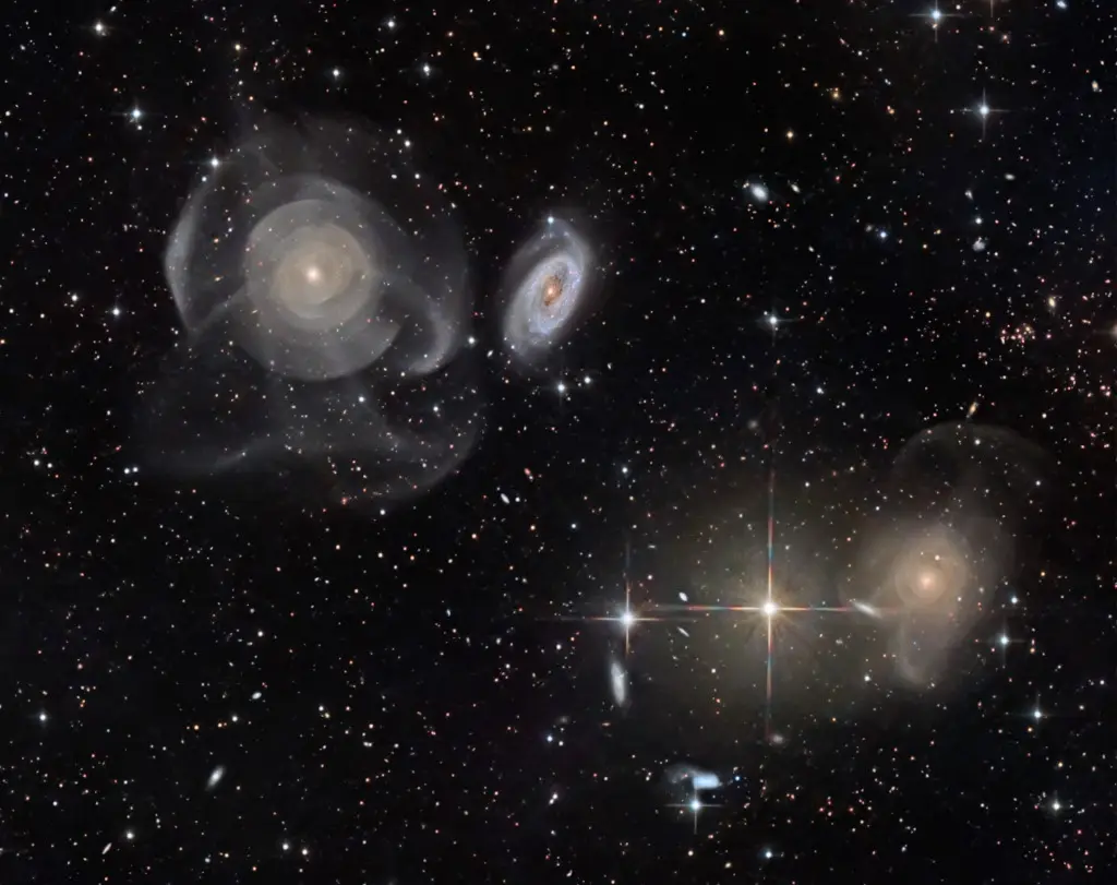 A deep space photograph shows several spiral galaxies and stars against a black background, with a prominent bright star in…