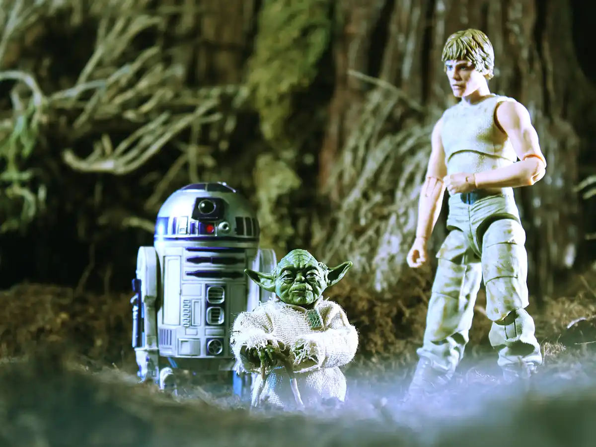 A staged photo of Star Wars action figures: Yoda, R2-D2, and Luke Skywalker standing in a forest-like setting. Yoda is in…